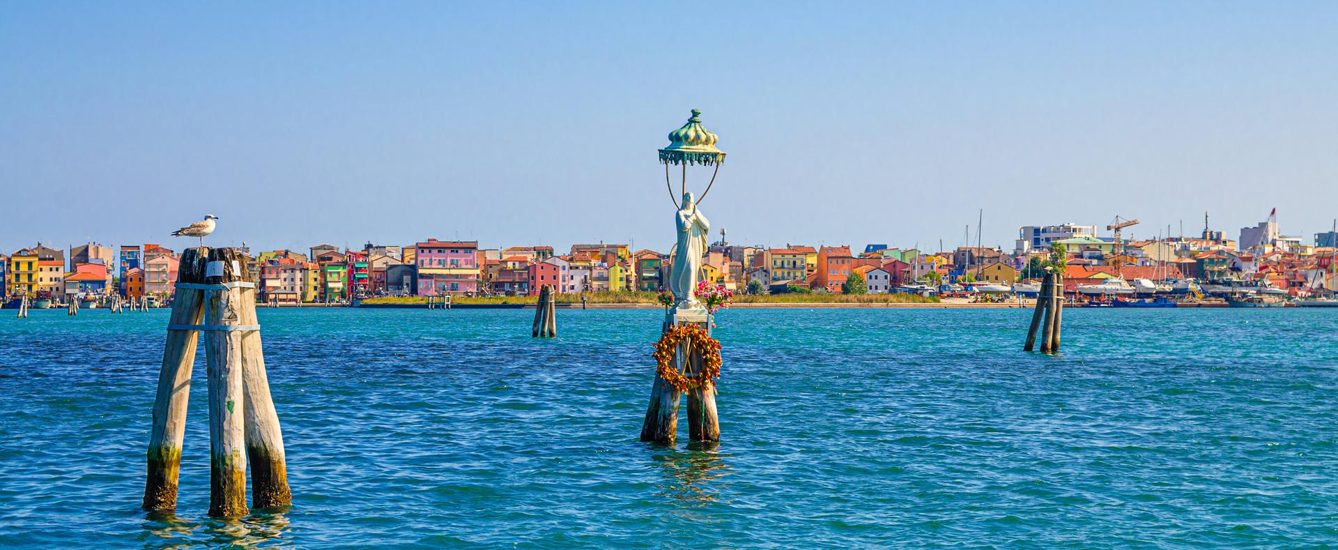Statua sull'acqua, case colorate, gabbiano su palo, atmosfera serena e vivace.