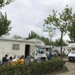 Camper parcheggiati, persone sedute all'aperto, alberi e cielo nuvoloso.