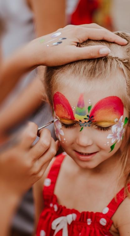 Bambina sorridente con trucco facciale colorato, indossa un vestito rosso a pois.