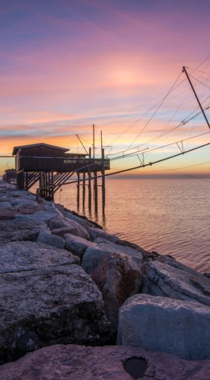 Tramonto su un trabucco sul mare, con cielo colorato e rocce in primo piano.