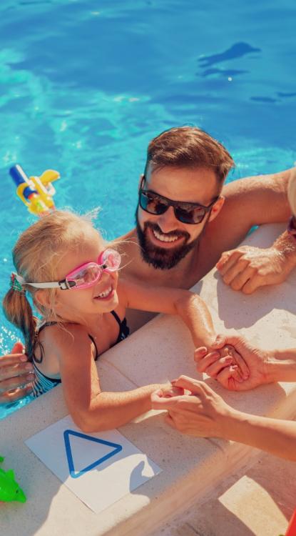 Bambina con occhialini in piscina, sorridente con adulto, vicino a un disegno triangolare.
