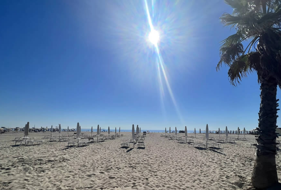 Sonniger Strand, geschlossene Sonnenschirme, klarer Himmel, entspannte Sommerstimmung.