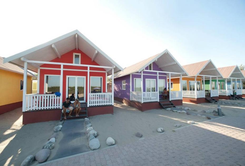 Case colorate con veranda su una spiaggia, circondate da pietre e sabbia.