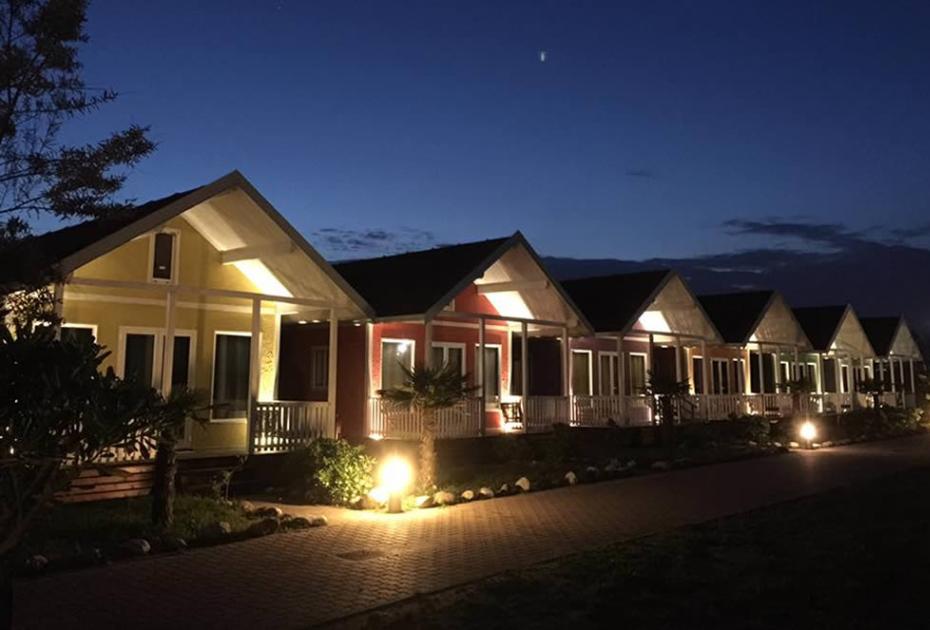Bungalow colorati illuminati di notte, lungo un viale alberato, sotto un cielo stellato.